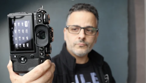 Man in glasses and a black jacket holding up a black mirrorless camera toward the viewer, its rear screen showing menu icons.