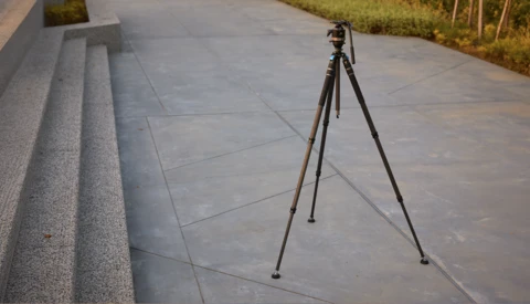 Dark tripod with a fluid video head and panning handle standing on a concrete plaza beside granite steps