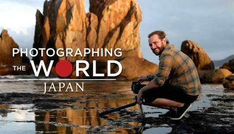 Photographer crouching at rocky coastline with camera, framed by golden sea stacks under dramatic sky