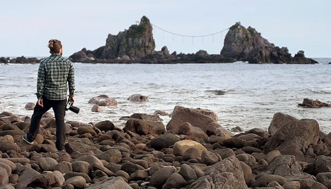 Photographer standing on rocky beach facing rocky islands connected by cables offshore.