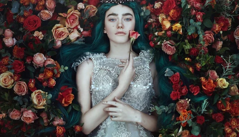 Woman with long dark hair and tiara wearing beaded silver gown, lying among vibrant red and orange roses.