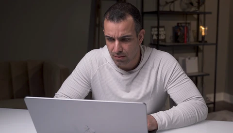 Man in light gray long-sleeve shirt focused on laptop screen at white desk.