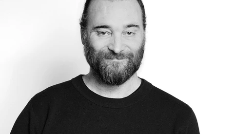 Black and white headshot of a bearded man with slicked-back dark hair wearing a black crew neck t-shirt.