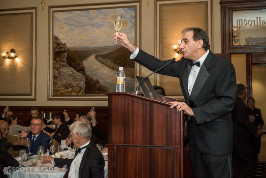 an event photo of a man holding his wine glass out from behind a podium, proposing a toast