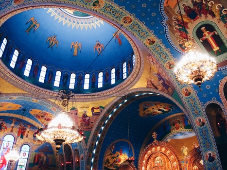 Interior of ornate Orthodox church with blue domed ceiling, golden religious iconography, and hanging chandeliers.