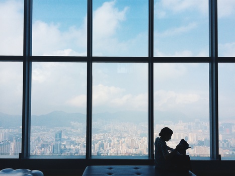 Silhouetted figure sitting against a large gridded window overlooking a sprawling cityscape.