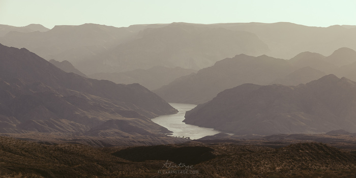 Layered mountain ranges with a winding river cutting through a desert valley under hazy atmospheric conditions.