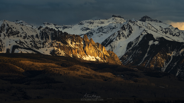 Snow-capped mountain range with golden-lit cliff face in dramatic cross-lighting beneath storm clouds.