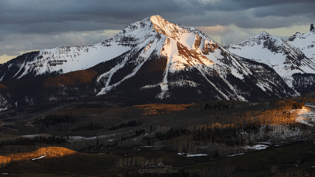 Snow-capped mountain peak illuminated by golden light during sunset, with forested slopes and valley below.