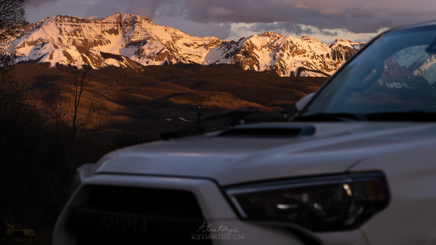 SUV hood and windshield in foreground with snow-capped mountains at golden hour in background.
