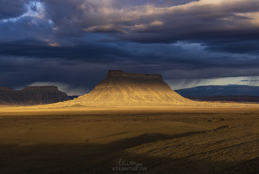 Flat desert landscape with a distinctive flat-topped mountain lit by golden sunlight under stormy clouds.
