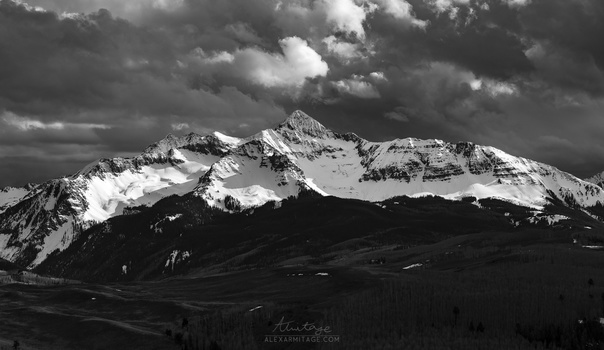 Snow-capped mountain peak with forested foothills under dramatic cloudy sky in black and white.