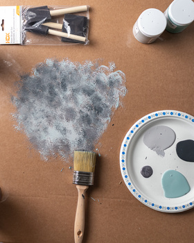 Paintbrush with wet gray paint on brown paper surrounded by art supplies and color palette.