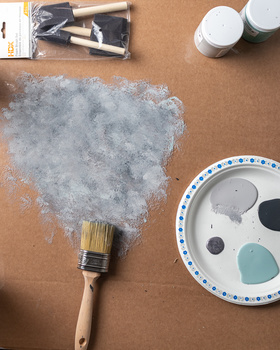 Paintbrush with gray paint swatch on kraft paper surrounded by art supplies and paint palette.