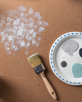 Paintbrush, scattered white paint, and palette with blue trim on tan surface.