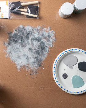 Artist's workspace showing a textured gray-blue paint swatch on kraft paper surrounded by brushes, paint containers, and a ceramic palette.