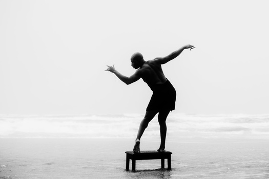 Silhouette of a person balancing on a platform in shallow water, arms extended in an expressive pose.