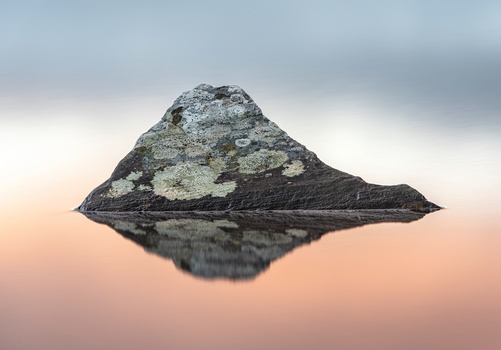 Rocky mountain peak with lichen-covered slopes reflected in still water at sunset.