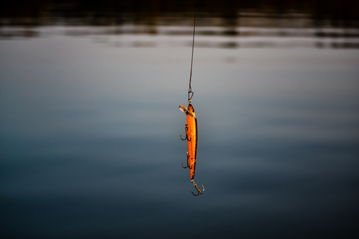Orange fishing lure suspended on a line above calm water with a blurred shoreline background.