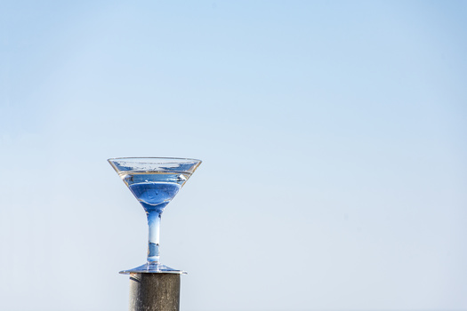 Blue cocktail in a martini glass balanced on a wooden post against a clear sky.