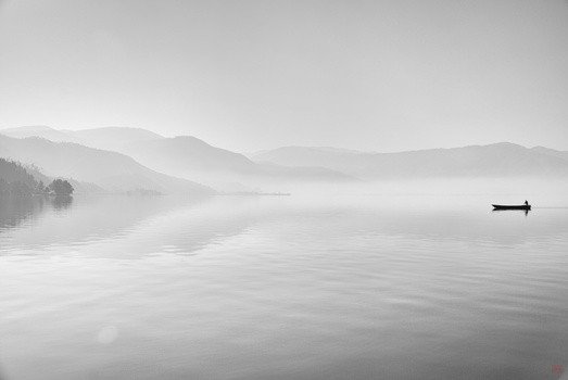 A solitary boat on calm, misty water with forested shoreline and mountains receding into fog.