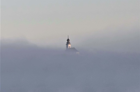Church steeple with illuminated spire emerging from dense fog during twilight hours.