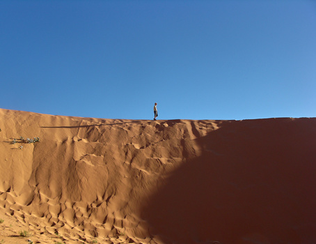 Lone figure standing on the crest of a sandy desert dune against clear blue sky.