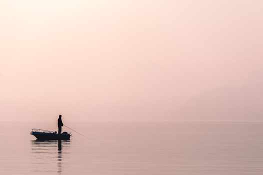 Silhouetted fisherman standing in small boat on perfectly still water at dawn, reflected below.