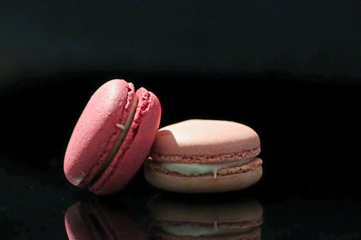 Pink and pale beige macarons arranged on a reflective dark surface with dramatic side lighting.
