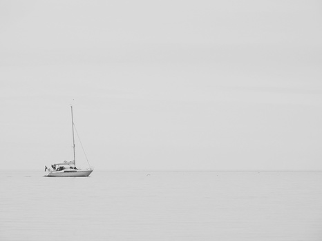 Solitary white sailboat anchored in calm, misty water under overcast sky.