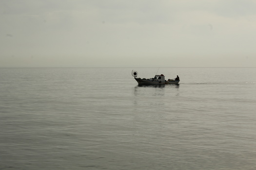 Small fishing boat on calm, misty water with minimal visibility and neutral gray tones.