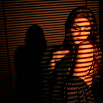 Figure backlit by warm orange light filtered through horizontal venetian blinds, creating striped shadow patterns across the body.