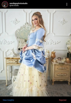 Woman in blue blouse and cream tulle skirt posing beside ornate furniture in classical interior.