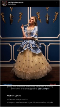 Woman in ornate ball gown with lace bodice and tulle skirt poses against blue wall with gold trim and decorative elements.