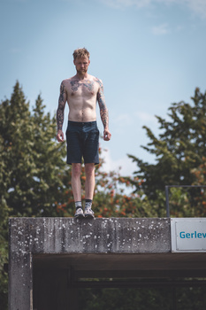 Shirtless male figure standing on concrete platform against blue sky with evergreen trees.