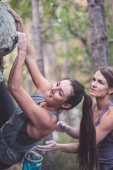 Two women spot a male climber as he reaches upward on an outdoor rock face.