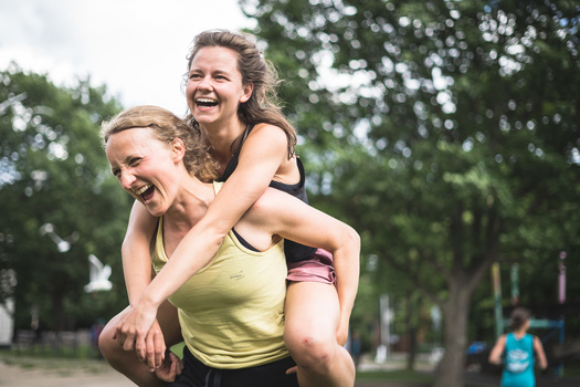 Woman in yellow tank top giving piggyback ride to smiling girl in park setting.
