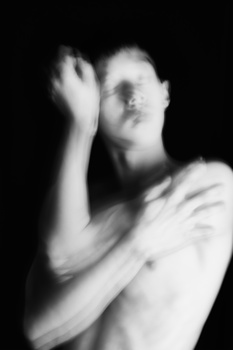 Shirtless person with hand raised to face, shot against black background in high-contrast black and white.