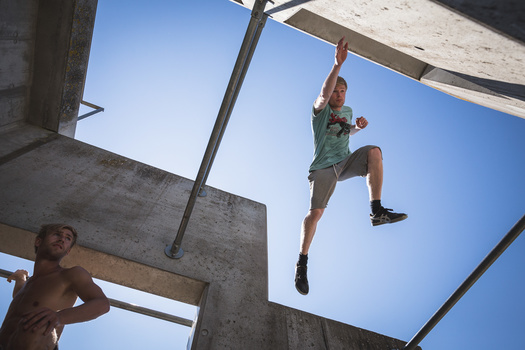Athlete suspended mid-air while climbing a concrete obstacle course structure against a clear blue sky.