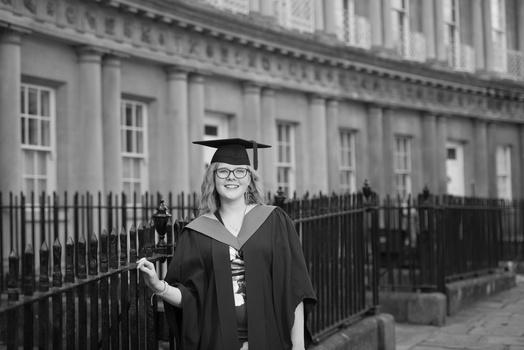 Graduate in cap and gown smiling at camera in front of classical Georgian architecture.