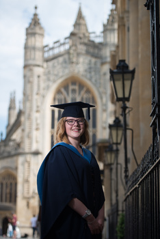 Graduate in mortarboard and academic gown poses in front of Gothic cathedral architecture.