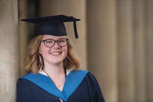 Graduate in academic regalia and glasses smiling at camera against neutral background.