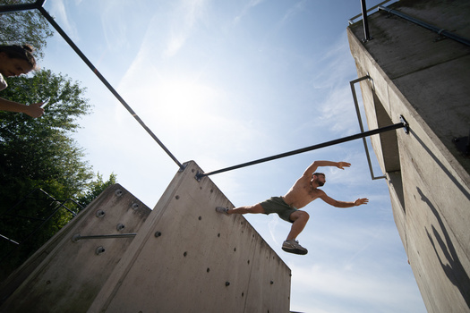 Athlete mid-leap between two concrete climbing walls under bright daylight.