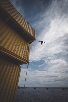 Person in mid-air diving or falling from stacked yellow shipping containers toward calm water below.