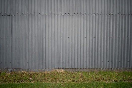 Corrugated metal wall with horizontal ridged texture and bolted seams, grass visible at base.