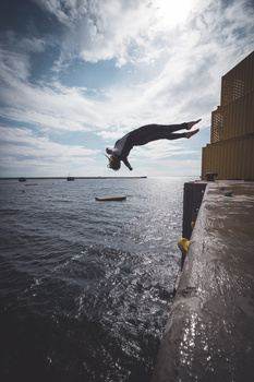 Person mid-dive off a wooden pier into calm water under a blue sky with white clouds.