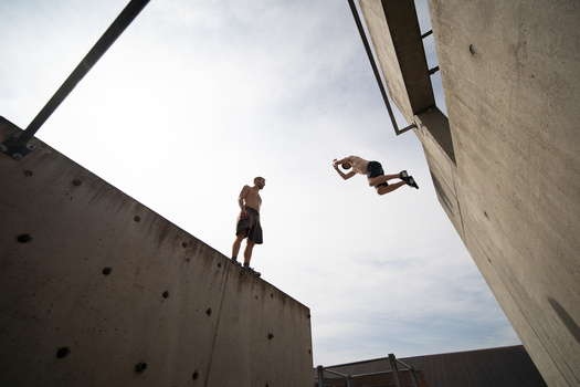 Two athletes performing parkour jumps between concrete walls in bright daylight.
