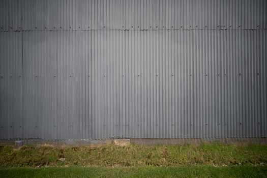 Corrugated metal wall with horizontal ribbing and rivets above overgrown grass and bare ground.