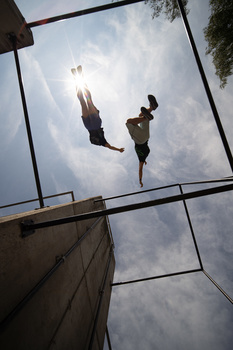 Two people jumping on a trampoline, photographed from below against a cloudy sky.