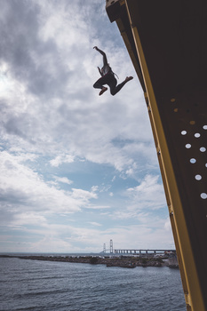 Person mid-air in freefall, shot from below against cloudy sky with industrial pier structure visible.